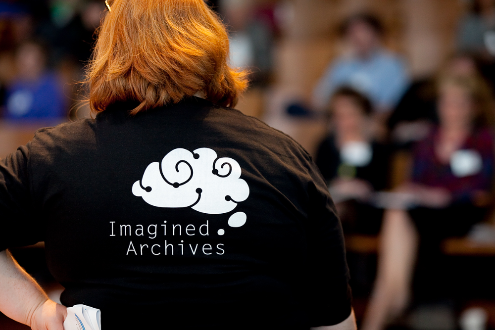 Person from behind facing audience; shirt shows brain‑cloud logo above 'Imagined Archives' text