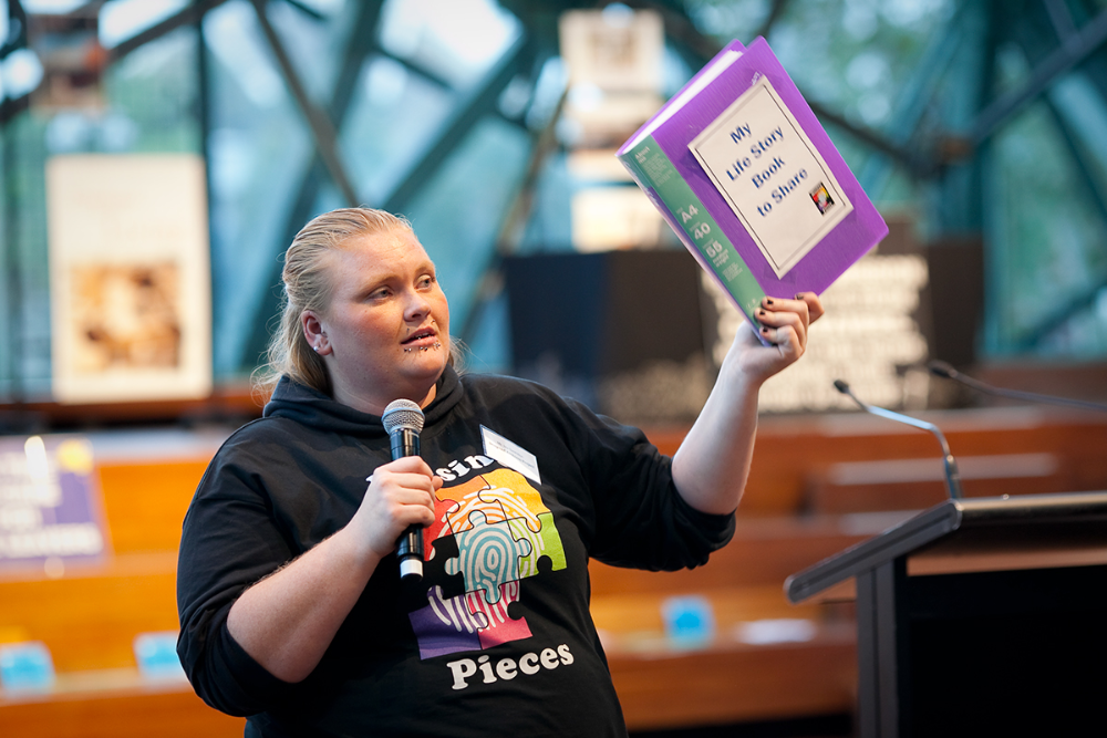 Presenter at 'Rights of the Child' summit holding 'My Life Story Book to Share' and microphone, wearing hoodie with thumbprint puzzle graphic and 'Missing Pieces' text.